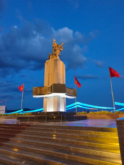 The image shows a monument in the city of An Nhơn, Vietnam, at dusk. The monument is a tall, stone structure topped with a sculpture of figures holding flags. Three red flags with a yellow star are visible, two on flagpoles near the monument and one to the left in the background. The monument is illuminated, and the surrounding area is lit with blue lights, possibly from a bridge or walkway in the background. Steps made of granite lead up to the monument, and the ground is wet, reflecting the lights. The sky is a deep blue with some clouds. There are no people visible in the image.