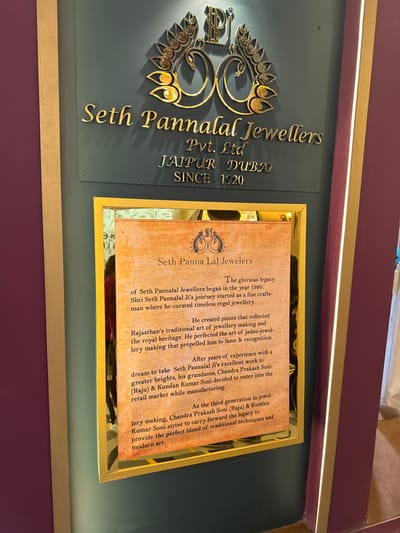 The image displays an interior wall feature for "Seth Pannalal Jewellers Pvt. Ltd." located in Jaipur, India, within the city of Jaipur Municipal Corporation. The upper section consists of a dark teal panel with raised, metallic gold lettering, stating the business name, its presence in "Jaipur Dubai," and its establishment "Since 1920." A prominent gold emblem, incorporating stylized "P" and "L" letters with intricate floral or peacock-like motifs, is centered above the text. Directly beneath this sign, a rectangular historical plaque is mounted. This plaque is framed in gold and covered by reflective glass, bearing the title "Seth Panna Lal Jewelers." The text, printed on a textured background resembling aged parchment, details the company's legacy, its 1920 founding by Shri Seth Pannalal Ji, and the continuation of "jadau-jewellery making" by his grandsons, Chandra Prakash Soni (Raju) and Kundan Kumar Soni. The immediate environment includes sections of maroon wall on the left and right periphery, a vertical golden trim strip on the left, and a vertical white light strip on the right. Reflections, likely of the photographer, are visible on the glass surface of the historical plaque. No other individuals or direct actions are present.