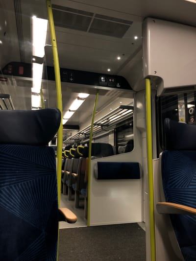 Inside a train car, navy blue patterned seats with armrests sit across from each other. A yellow bar runs horizontally along the sides, serving as a handrail. White walls with small circular lights line the interior. A section of the wall is a dark gray color and displays white icons and arrows. Small pull-down trays, some with blue cushions, are attached to the back of the seats. The aisle is covered in gray carpeting.