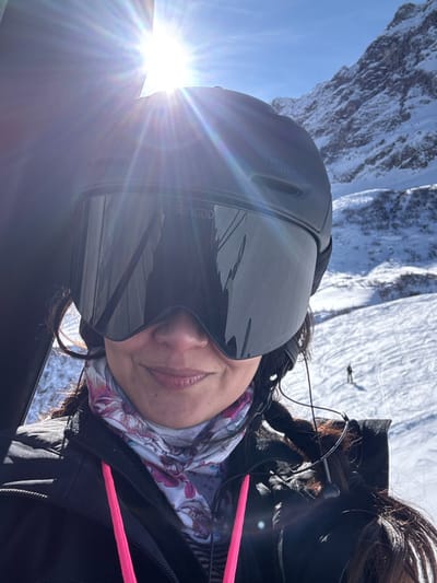 The image displays a close-up, selfie-style photograph of a female individual against a snow-covered alpine environment in Zürs, Austria, under a clear, bright blue sky. The individual is centered in the foreground, wearing a matte black ski helmet with a visible "MIPS" logo, integrated with oversized, dark-tinted ski goggles that display "SUNGOD" on the upper left lens and "KOSMOS" on the lower right. The goggles obscure the upper half of her face; her lower nose, mouth, and chin are visible, showing a slight smile. She wears a white neck gaiter with a pink and purple floral pattern, and a black ski jacket, revealing a striped garment underneath. Bright pink straps or cords are present on her chest area. Black wired earbuds are visible, with a cable extending from her right ear. Her dark hair is visible around her ears and shoulders. The background consists of expansive snow-covered mountains and slopes. A strong sunburst and lens flare are prominent in the upper left quadrant of the image. A small, distant figure, identified as a skier, is visible on the lower right section of the snow-covered slope.