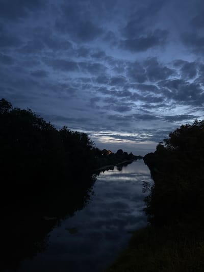 The image presents a wide-angle, low-light outdoor scene of a waterway under an overcast sky, consistent with either dusk or dawn in Boston, United Kingdom.

The upper approximately 40% of the frame is dominated by a dense formation of dark grey to muted blue stratocumulus or altocumulus clouds, exhibiting varied textures and shading. A narrow, lighter band of sky is visible along the horizon line above the distant landscape, indicating residual ambient light.

A calm waterway, likely a canal or river, occupies the central portion of the image, extending from the foreground into the deep background. The water's surface reflects the overhead cloud cover, displaying dark grey and bluish tones with indistinct cloud patterns. Scattered patches of aquatic vegetation, possibly water lilies or algae, are visible on the water, particularly in the lower-left quadrant.

Both banks of the waterway are densely lined with dark, silhouetted foliage, primarily trees and shrubs. The vegetation on the left bank is more prominent and appears to consist of taller deciduous trees. In the immediate lower right foreground, some darker, indistinct grass or reed-like vegetation is present on the bank.

On the left bank, in the mid-distance, a residential-style building with a reddish-brown pitched roof and lighter-colored walls (suggestive of brick) is partially visible. Further along the same bank, a second, less distinct structure can be observed. In the far distance, spanning the waterway, a dark, metallic truss bridge structure is faintly discernible against the lighter horizon.

No individuals or direct actions are depicted. The ambient lighting suggests a time of day with very low natural light. The presence of a canal or river, residential buildings, and a bridge is consistent with the urban and semi-rural landscape of Boston, Lincolnshire, United Kingdom, which features waterways like the River Witham and Maud Foster Drain. The architectural style of the visible building also aligns with typical UK construction.