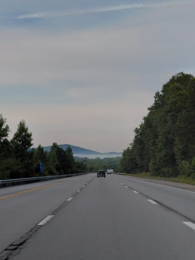 This is a sideways, high-angle shot of a highway in Berea, United States. The road appears to be in a rural or semi-rural area, bordered by dense trees on the left and a lighter, hazy sky with some wispy clouds on the right. In the distance, rolling hills are visible, shrouded in a light mist, suggesting either early morning or a generally overcast day. Two vehicles are on the road: a white SUV and a black vehicle behind it, both moving away from the viewer. The road itself is asphalt with distinct white lane markers and a yellow dividing line. There are also guardrails along the side of the road. The overall impression is one of travel through a natural landscape.