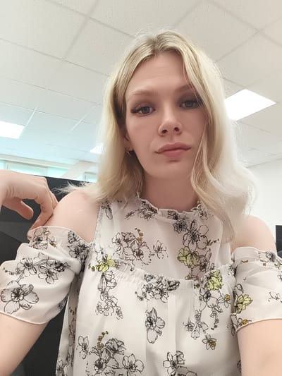 This is a portrait-style photograph of a young woman with blonde hair and light blue eyes, looking directly at the camera. She is wearing a white off-the-shoulder top with a floral print. Her left arm is bent, and her hand is resting on her shoulder. The background appears to be an indoor setting with a dropped ceiling and fluorescent lighting, possibly an office or classroom. There is no discernible information within the image that places the location in Postol, Russia.