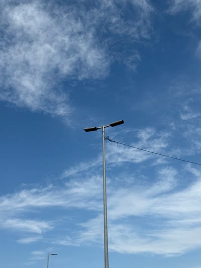 A tall, silver-colored street lamp with two lights is positioned in the lower half of the frame, angled slightly to the left. A thin, black wire extends horizontally from the lamp, disappearing to the right. The lamp is set against a bright blue sky with wispy, white clouds scattered across it. In the far lower left corner, a portion of another, shorter street lamp is visible. The image appears to be taken outdoors, in an open area within the city of Volkhov, Russia.