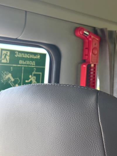 This media file features a close-up view of a seat in what appears to be a public transportation vehicle, possibly a train or bus, given the presence of an emergency exit sign and a red safety tool. The seat is upholstered in textured gray material, with visible stitching along the edge.
In the background, a bright red object, likely a safety hammer or similar emergency tool, is mounted on a vertical surface. It has a distinctive shape with a handle and a striking end.
To the left of the seat, a green emergency exit sign is partially visible through a window or opening. The sign displays pictograms and text in Russian. The visible text reads "Запасный" and "Выход," which translates to "Emergency" and "Exit" respectively. Pictograms depict figures in motion, suggesting evacuation instructions.
The lighting suggests an indoor setting, possibly during daytime, as there is ambient light illuminating the scene. The overall impression is one of preparedness for emergencies within a transportation context. No people are directly visible in the frame. The setting appears to be a clean and functional interior. The location context provided, Мастоцкі сельскі Савет, Belarus, indicates a rural administrative region in Belarus, which could imply the transportation might be local or regional.