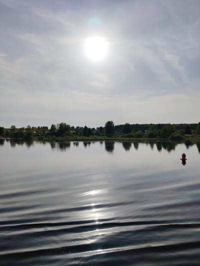 The image depicts a body of water under a bright, cloudy sky with the sun prominently visible. A line of trees and vegetation is visible on the far shore. A red navigational buoy is located in the water on the right side of the image. The water has gentle ripples that reflect the sunlight. The location is identified as Irdomatka, Russia.