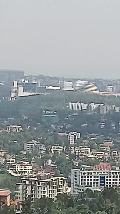 This image presents an elevated, long-distance view of a densely developed urban landscape in Kigali City, Rwanda, under an overcast sky. The scene depicts numerous multi-story buildings interspersed with extensive green vegetation covering rolling hills.
In the mid-ground, a prominent, well-treed hill features several large, modern structures. To the right, the distinctive white, curved roof and facade of the Kigali Convention Centre are clearly visible. Adjacent to it, to the left, is a complex of modern, multi-story buildings, including the Radisson Blu Hotel. Further to the left on the same hill, another multi-story structure with a segmented facade is present. In the far upper-left background, partially obscured by atmospheric haze, the outline of a large stadium, identifiable as the BK Arena, is discernable.
Below these prominent landmarks, in the foreground and mid-ground, numerous residential and commercial buildings populate the hillsides. These buildings vary in height, typically ranging from three to eight stories, and exhibit diverse architectural styles with facades in shades of white, grey, and brown, some featuring blue window accents or reddish-brown tiled roofs. A large building in the lower mid-right has a prominent white facade with numerous square windows and some blue-paneled sections. Another multi-story building in the lower-left features a light-colored facade with many windows and what appears to be a flat roof with dark elements. One structure in the mid-left appears to be under construction or undergoing renovation, partially covered by a light-green tarp or netting. No individuals or specific actions are discernible due to the distance and resolution. The overall environment suggests a bustling urban area integrated with its natural, hilly topography.