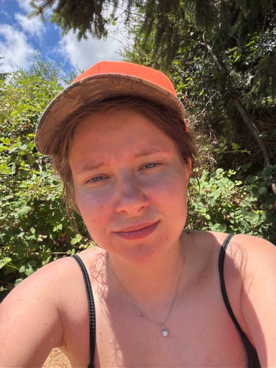 A young woman, appearing to be in her late teens or early twenties, is pictured outdoors, possibly in Cherry Grove, United States, given the hint. She is wearing an orange baseball cap, a black tank top, and a silver necklace with a small pendant. Her skin appears to be wet, with visible water droplets on her chest and arms, suggesting she may have been swimming or is in a humid environment. The background consists of lush green foliage and trees, with a glimpse of a blue sky with white clouds overhead. The lighting indicates it is daytime.