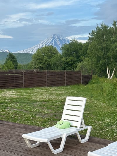 In Yuzhnyye Koryaki, Russia, a white plastic chaise lounge sits on a wooden deck. A green towel is draped over the lounge. Beyond the deck, a well-maintained lawn is dotted with small white flowers. A dark wooden fence runs horizontally across the middle ground, separating the lawn from a dense forest. In the background, a snow-capped volcano dominates the horizon under a cloudy sky. The environment appears serene and natural, with a hint of recreational setup.