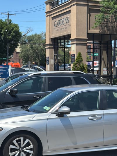 The image depicts an exterior scene in the Village of Great Neck Plaza, United States of America. In the foreground, a silver sedan is parked, its front wheel and driver's side visible. The driver, a young person with dark hair, is looking forward through the windshield. Behind the sedan, a black SUV is parked, partially obscuring the silver car. Further back, a multi-story building with "THE GARDENS AT GREAT NECK PLAZA" signage is visible, with a large arched entrance. Several other vehicles and trees are present in the background. The sky is clear and blue, suggesting a daytime scene.