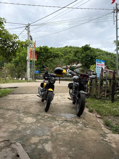 Two motorcycles, one yellow and one dark gray, are parked on a paved surface with a slight incline. The yellow motorcycle is on the left, facing slightly to the right, with a black helmet resting on its rear. The dark gray motorcycle is on the right, facing slightly to the left, with a black bag attached to its handlebars.
In the background, there is a wooden fence, a building with a thatched roof, and a lush green hillside covered in trees. A blue and white sign with Vietnamese text is visible to the right of the motorcycles. A utility pole with numerous wires is located to the left of the scene. Red and yellow flags are also visible on a pole near the utility pole. The ground is a mixture of concrete and patches of dirt and grass. The sky is overcast. The scene appears to be in a rural or semi-rural area. The sign indicates the location is within Kim Ngân Commune, Vietnam.