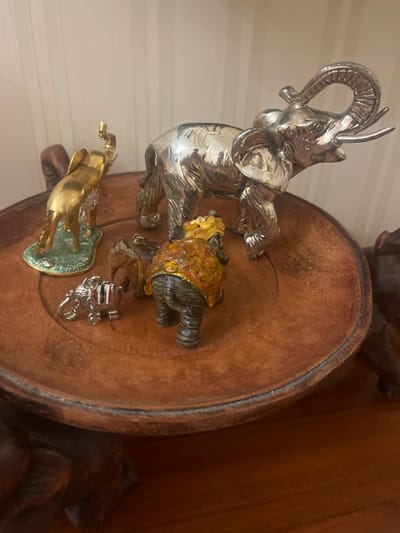 The image shows a collection of decorative figurines placed on a wooden surface. The prominent subjects include a shiny silver elephant figurine with its trunk raised, a smaller gold-colored horse figurine on a green base, and a unique figurine that appears to be a turtle or tortoise embellished with amber-colored stones. There's also a small silver charm with red and blue stripes. The figurines are arranged on a circular wooden platter, which itself rests on a darker wooden surface. The background is out of focus but suggests an indoor setting with vertical white paneling on the left and a wooden surface on the right. There is no discernible activity or event taking place, and the scene is static, suggesting a display of ornamental items. Notable details include the reflective quality of the silver elephant and the textured appearance of the amber on the turtle figurine. No text is visible in the image. The location context provided is Edem, Russia, but there are no visual cues within the image to confirm this location.