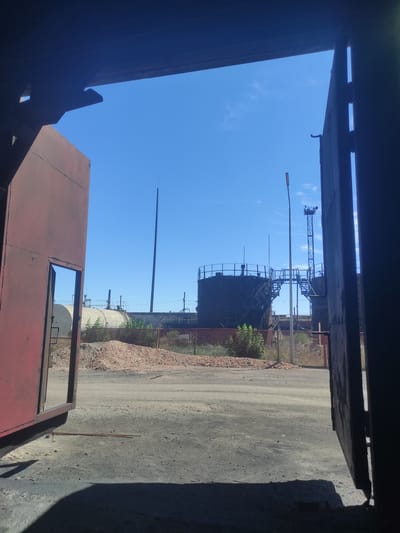 The image captures a view looking out from a dark, partially open structure onto an industrial area under a clear blue sky. In the foreground, the edge of a large, weathered red metal structure is visible on the left, and the edge of a dark, vertical structure is on the right, framing the scene. The ground is a dusty, gravelly surface with some scattered debris.
Beyond the immediate foreground, the industrial landscape unfolds. Two large, dark, cylindrical storage tanks with elevated walkways and staircases are prominent in the mid-ground. To the left of these tanks, a long, lighter-colored cylindrical storage tank is partially visible, running horizontally. A tall, slender antenna or tower rises into the sky behind the tanks. Several smaller structures, pipes, and industrial equipment are scattered throughout the area. A fence separates the industrial tanks from a large pile of reddish-brown material, possibly slag or coal, to the left of the tanks. Green foliage is visible around the base of the tanks and behind the fence. A street lamp is positioned between the foreground and the storage tanks.
The scene suggests a day in an industrial zone, possibly a processing plant or storage facility. The lighting indicates it is daytime with bright sunlight. There are no visible people in the image. The location appears to be an industrial site in Kyzyl, Kazakhstan, given the context provided.