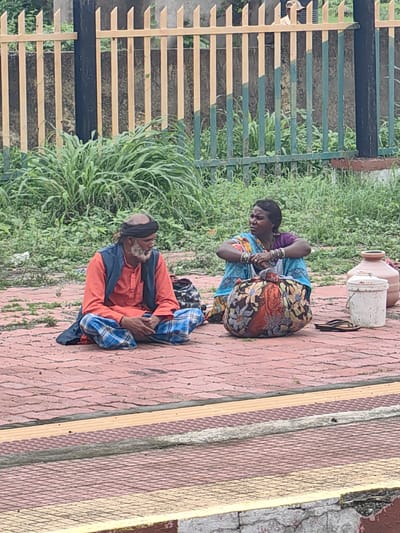 In Shihar, India, on a paved area with a yellow-lined border, two individuals are seated on the ground amidst overgrown vegetation and a metal fence. A man, wearing a black turban, an orange kurta, a dark vest, and plaid pants, sits on the left. To his right, a woman, adorned with bangles and wearing a colorful patterned saree over a purple blouse, is seated. In front of her is a large, floral-patterned bag. Nearby, a white bucket and two clay pots are visible. The scene appears to be outdoors, possibly near a railway platform or a public gathering space.