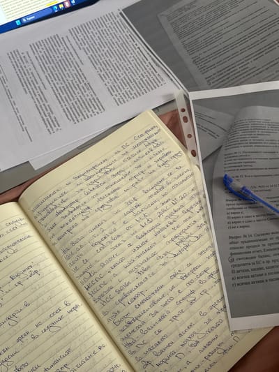The image captures an overhead view of a study or work desk, featuring several printed documents, an open notebook, and a blue ballpoint pen. The setting appears to be indoors, well-lit, suggesting daytime.
The main subjects are various papers and a notebook, indicating an activity involving reading, learning, or working. One document is folded and partially obscures another, while a third is open and covered with handwritten notes in a spiral-bound notebook. A blue pen rests on one of the printed pages.
The printed documents contain text primarily in Bulgarian Cyrillic script, with titles and questions related to accounting, finance, and the public sector. Visible text includes phrases like "Въпрос №3. При подготовката на финансовите отчети..." (Question #3. When preparing financial statements...), "МССПС 2 Отчет за паричните потоци" (IPSAS 2 Statement of Cash Flows), "Въпрос № 11. Последваното оценяване и публичния сектор е:" (Question #11. Subsequent measurement and public sector is:), and mentions of "финансовото състояние" (financial position), "публичния сектор" (public sector), "активи и пасиви" (assets and liabilities), and "бюджет" (budget). One document explicitly references "Handbook of International Public Sector Accounting Pronouncements."
The handwritten notes in the notebook are also in Bulgarian Cyrillic, discussing topics such as "Нормативен подход" (Normative approach), "Бюджетирането на ПС" (Budgeting of the Public Sector), "финансите" (finance), "счетоводния" (accounting), "статистиката" (statistics), and references to "МССПС" (IPSAS) and "НСС" (National Accounting Standards). A partially visible hand, possibly belonging to the note-taker, is seen holding the notebook.
The overall scene depicts an academic or professional environment focused on financial or public sector accounting studies in Mechkarevo, Bulgaria.