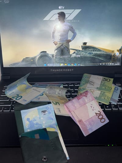 A low-angle, medium shot captures a dark-colored laptop with an "F1: THE MOVIE" wallpaper on its screen. Scattered across the keyboard and extending onto the screen area are several banknotes, appearing to be both Euros and Belarusian Rubles. A dark green, small wallet or cardholder is partially visible on the keyboard, containing a blue card and Euro banknotes. The environment is indoors, with the laptop being the central subject. The overall lighting suggests an indoor setting, possibly a home or office. No people are visible in the image, and no specific city or location like Kobryn, Belarus, is identifiable from the visual cues.