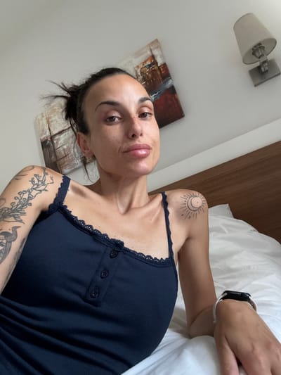 A selfie taken from a slightly high angle shows a woman lying on a bed. She is wearing a dark blue tank top with lace trim and three buttons down the front. She has several tattoos on her left arm and a small tattoo of a sun and moon on her right shoulder. She is wearing a white watch on her left wrist. Her hair is pulled back in a messy bun. She has freckles on her face and her lips are slightly parted. Behind her, a wooden headboard of a bed is visible, along with a white duvet. On the wall behind her, there are two abstract paintings and a wall-mounted lamp. The scene appears to be an indoor setting, possibly a bedroom. There is no specific indication of the location being the city of Aksu, Turkey.