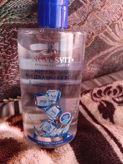 The image shows a bottle of "NOVOSVIT" micellar water resting on a textured fabric surface. The bottle is transparent, revealing the clear liquid inside. The label on the bottle features text in Russian, including the brand name "NOVOSVIT," "PHARMA LABORATORY," and "МИНЕРАЛЬНАЯ МИЦЕЛЛЯРНАЯ ВОДА" (Mineral Micellar Water). It also indicates that the product "ОЧИЩАЕТ + СНИМАЕТ МАКИЯЖ, ВОССТАНАВАЛИВАЕТ + УСПОКАИВАЕТ" (Cleanses + Removes Makeup, Restores + Soothes). Small graphics of ice cubes with chemical symbols "Mg," "Na," and "Mn" are visible on the label, along with a circular blue emblem that reads "БЕЗ ПАРАБЕНОВ И ОТДУШКИ" (Without Parabens and Fragrance). The bottom of the label has text that appears to say "ДЛЯ ЛИЦА, ГУБ И ГЛАЗ" (For Face, Lips, and Eyes). The background fabric is a blend of colors, predominantly brown and beige, with a striped pattern. There is no discernible indication of the location being the city of Monastirische, Russia, as the image only depicts the product and its immediate surroundings. There are no people or any actions depicted in the image.
