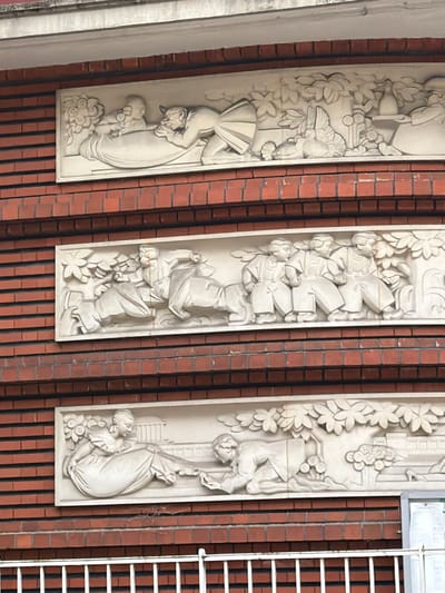 The image shows three vertical bas-relief panels set against a brick wall. Each panel depicts a scene with stylized figures and foliage. The leftmost panel appears to show a woman in a flowing garment holding a baby or a small child. The middle panel features several figures in what seem to be traditional clothing, possibly a group of children or workers, with their arms linked or reaching out. The rightmost panel depicts a figure in a dynamic pose, potentially falling or reaching, with a flowing garment and a smaller figure or object nearby.
The style of the reliefs is classical, with a focus on flowing lines and expressive forms. The foliage and floral elements suggest a connection to nature or a garden setting. The figures' expressions are not clearly discernible due to the resolution and lighting, but the overall impression is one of movement and perhaps communal activity or narrative storytelling. The setting appears to be an exterior wall, possibly part of a building's facade, with red brickwork providing a textured background. The lighting suggests it is daytime, though the weather is not evident. There is no visible text in the image.