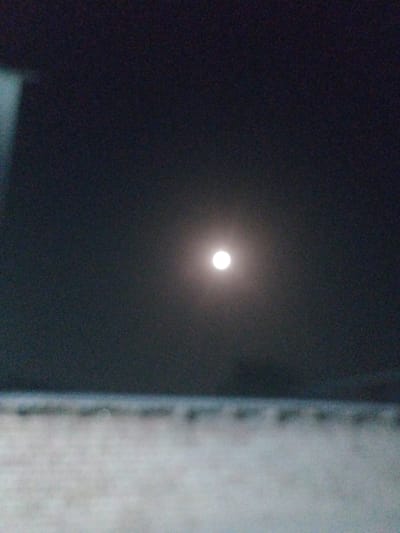 The image depicts a night scene with a full moon prominently featured in the dark sky. The moon is a bright, circular object, slightly diffused by the atmosphere. The sky is a deep, dark gray, suggesting it is nighttime. In the foreground, there is a blurry, horizontal band of lighter color, possibly indicating a surface like a building or ground, but it is out of focus and not clearly defined. There are no people or any discernible activity visible in the scene. The image appears to have been taken from a low vantage point. The location is identified as Социализм, Kyrgyzstan.