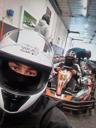 This image depicts a go-karting track in Maasvlakte, Rotterdam, Netherlands. In the foreground, a person wearing a white and black helmet, a black balaclava, and a dark jacket is captured in a selfie. Their eyes are visible through the helmet's opening. Behind them, another individual, also wearing a helmet and a dark jacket with white stripes, is seated in a go-kart with the number "35" visible on its front. This individual is holding the steering wheel. A third person in a dark hooded garment is leaning over the go-kart. The environment appears to be an indoor facility with multiple go-karts parked in rows, characteristic of a go-karting arena. There are also some industrial-looking elements and a poster of a race car on the wall.