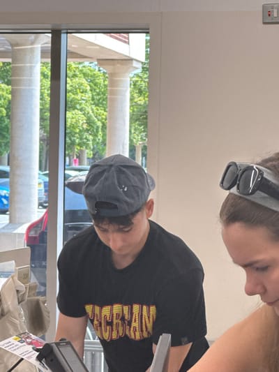 Two people are captured in a slightly skewed angle. One, partially visible at the top, has sunglasses on their head. The other wears a dark t-shirt and a baseball cap, focused on something near a cash register. Outside the window behind, cylindrical pillars frame a view of green trees and glimpses of cars in the distance.