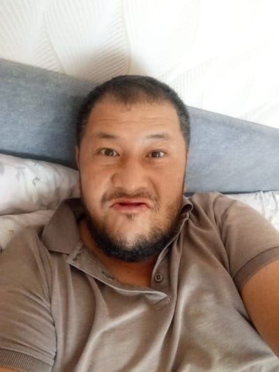 A close-up, selfie-style photograph shows a man with dark hair and a beard lying on a bed. He is wearing a tan polo shirt. His head is slightly tilted, and he is looking directly at the camera with a neutral expression. The background consists of a grey headboard and a white, textured wall. The lighting is bright, suggesting it is daytime.