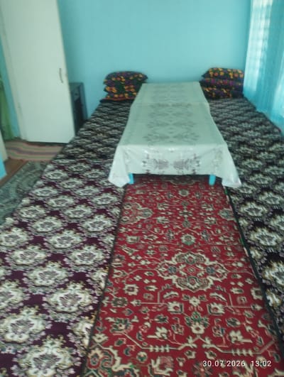 The image shows a room with blue walls and floor coverings. A white table with a patterned tablecloth is placed on a floor covered with several carpets. One carpet is red with intricate patterns, another is dark with a white floral design, and a third is a striped rug. There are also pillows with a floral pattern stacked on one side of the room. The room appears to be in a home or guesthouse in Quyosh, Uzbekistan, likely a living area or bedroom. The lighting suggests it is daytime.