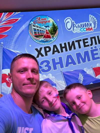 In Krasnyi Vostok, Russia, a man and two young boys pose for a selfie. The man, wearing a blue t-shirt, is in the foreground, smiling. To his right, two boys are close together, also smiling. One boy has red and blue face paint on his cheek. Behind them, a large screen displays a logo with text that reads "Алые Паруса" (Scarlet Sails) and "Орлята России" (Eaglets of Russia), along with the words "ХРАНИТЕЛИ ЗНАМЁН" (Guardians of the Banners). The screen also features images of a building, a sailboat, trees, and flags. The overall setting appears to be an indoor event or gathering.
