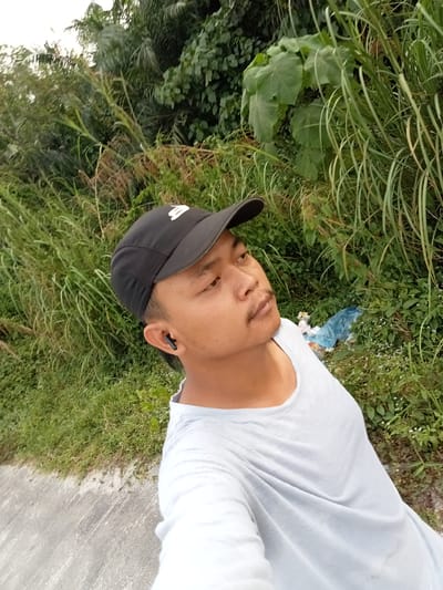 The image shows a young man wearing a black baseball cap, a light blue t-shirt, and earbuds, taking a selfie outdoors. He has a thin mustache. He is standing next to a road with tall green grass and lush vegetation behind him. The vegetation includes large-leafed plants and reedy grasses. Some blue plastic is visible among the plants in the background, suggesting litter. The lighting indicates it is daytime, possibly late afternoon or early morning due to the soft light and shadows. The overall impression is of a casual, outdoor setting likely in a rural or semi-rural area near Teluk Kenidai, Indonesia, given the vegetation and the apparent climate.
