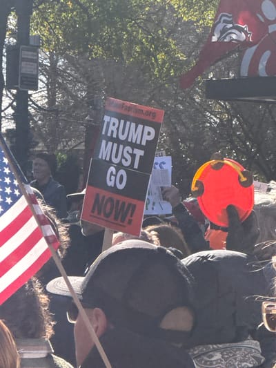 A political rally or protest scene shows a "Trump Must Go Now!" sign. The image includes a person holding an orange tambourine. An American flag is visible in the lower center. The protest appears to be outdoors. Other signs and people are present, suggesting a gathering of some size.