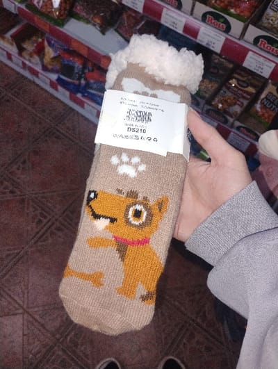 A human hand, partially obscured by a grey long-sleeved garment, holds a pair of beige knitted socks vertically in an indoor retail environment. The socks feature a cartoon design of an orange and brown dog with a pink collar and a white bone, and a white paw print on the sole. The sock cuffs are white and fuzzy. A white product tag, displaying "DS210", "MADE IN P.R.C.", a barcode, and material composition information (e.g., "82% acrylic, 17% polyester, 1% elastane"), is attached to the socks.

The background shows multiple tiers of retail shelving stocked with various packaged food items, predominantly nuts under the "Rois" brand. Some shelf labels display prices, such as "1.00 BGN." The flooring consists of dark reddish-brown patterned tiles. The exact location within the city of Byal Izvor, Bulgaria, is not identifiable from the visual information provided.