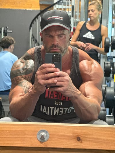 The image shows a muscular man taking a selfie in a gym. He is wearing a black tank top and a baseball cap. His right arm is heavily tattooed. In the background, a woman wearing an Adidas tank top is visible, as well as various weightlifting equipment. The location is not discernible from the image and there is nothing to indicate it is in Jemelle, Belgium.
