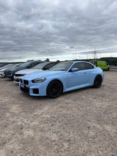 The image depicts a light blue BMW M2 (G87) coupe, featuring a black roof and black multi-spoke wheels, parked on an unpaved gravel surface under a heavily overcast sky. The scene is located in Hermsdorf, Germany.

The primary subject vehicle is positioned diagonally from the lower left to the upper right. Behind the BMW, a white car displaying a Swiss license plate "ZH 123 185" is visible, followed by a dark grey SUV with a partially visible German license plate (e.g., "SH GT 5398"), and other vehicles receding into the background. To the far right, a bright lime green van is parked. The ground consists of light brown and grey gravel and dirt.

In the mid-ground and background, a line of trees separates the parking area from the distant horizon. Several large construction cranes and multiple wind turbines are visible on the horizon, against the uniform grey sky. No individuals are present in the scene, and no actions or interactions are depicted.