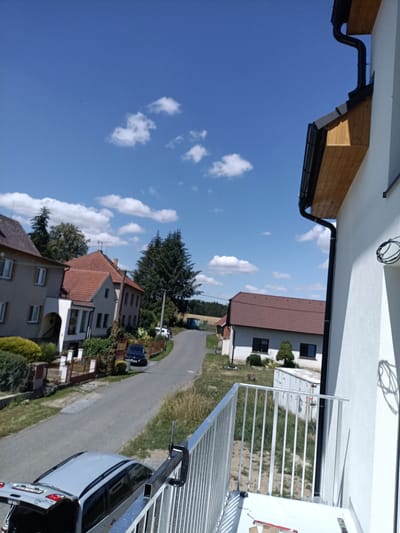 The image captures a sunny day in Rataje, Czechia, looking down a residential street from a balcony. On the left, two houses are visible. The closer house has a grey facade with white trim around its windows and a prominent white arched entrance. The second house, further down the street, has a terracotta tiled roof and a similar facade. A narrow asphalt road curves away from the viewer, bordered by grass and a few trees. A dark blue car is parked on the right side of the road. In the foreground, the edge of a modern white building with a white metal railing is visible, suggesting the vantage point is from a balcony or upper-floor window. A light-colored car with its trunk open is parked in front of this building, partially obscuring a metallic clamp attached to the railing. The sky is a clear blue with scattered white clouds.