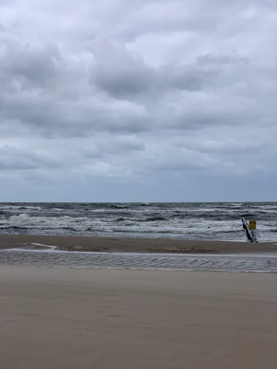 The image depicts a turbulent sea and beach scene, likely in Pobierowo, Poland. The sky is overcast with heavy, grey clouds that obscure the sun, creating a somber atmosphere. The sea is rough, with large waves crashing onto the shore, indicated by white foam and spray. A wooden structure, possibly a pier or breakwater, extends into the water from the right side of the frame. A yellow warning sign is affixed to this structure. A small, red buoy is visible in the sea near the wooden structure. The beach in the foreground is sandy and appears wet in places, with shallow pools of water reflecting the cloudy sky. The overall impression is one of a stormy or windswept day at the coast.
