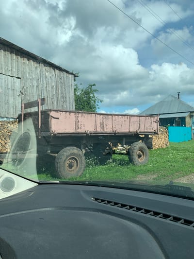 This is a medium-angle, outdoor shot of a dilapidated trailer parked in front of a wooden structure on a cloudy day. The trailer, with peeling pink paint, has two large, worn tires and a metal bed. To the left of the trailer, a stack of firewood is visible against the wooden building. Beyond the trailer, a blue structure and a grey-roofed building are present. The ground is covered in grass and some wildflowers, with a dirt track nearby. The image appears to be taken from inside a vehicle, with a portion of the dashboard and a speaker visible in the foreground. The scene is set in the rural environment of Barsai, Russia.