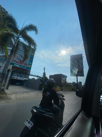 A sunny, clear sky illuminates a scene in Kertak Hanyar II, Indonesia. A person on a scooter, wearing a black helmet and jacket, rides down a paved road. The scooter has a white license plate with black text that reads "DA 8035 KF". On the left side of the road, a tall building with a prominent "YNL Stationery" sign and a palm tree stand against the blue sky. To the right, a large billboard displays various advertisements, with the sun glinting brightly behind it. The road is lined with power lines and a few other structures in the background, including a parked white car visible in an open garage. The image is taken from the perspective of a passenger inside a vehicle, with a portion of the car's interior visible in the foreground on the right.