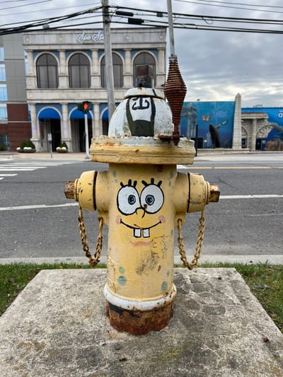 A yellow fire hydrant painted with a Spongebob Squarepants face sits on a concrete block by the roadside. It has chains connecting the caps and visible rust. Behind the hydrant is a street, and in the background are buildings, including one with a mural and another labeled "Sea Sta." A traffic light is visible near the entrance of the "Sea Sta" building, and there are power lines overhead. The sky is overcast.