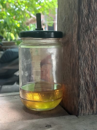 The image shows a glass jar filled with yellow liquid, possibly oil or fuel, resting on a weathered wooden surface. The jar has a black cap with a threaded bolt protruding from it. The background is blurred, with hints of green foliage suggesting an outdoor setting with trees and leaves. There are also some indistinct shapes in the background that could be furniture or other objects. The lighting suggests it is daytime, and the overall mood is neutral, focusing on the object itself. No legible text is visible on the jar or its surroundings. The scene appears to be a still life, with the jar as the central subject, possibly in a workshop or storage area given the wooden elements and the utilitarian nature of the object. The location context indicates Blangcot Tunong, Indonesia, which might imply a tropical or subtropical environment given the greenery.