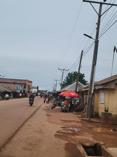 This image depicts a busy street scene in Tangu, Nigeria, likely during daylight hours with an overcast sky. The foreground features a dirt road with some puddles, suggesting recent rain or a naturally damp surface. On the right side of the road, there are several buildings with worn facades, some appearing to be small shops or market stalls. One stall has a prominent red umbrella with white lettering that is difficult to decipher.

The street is populated with numerous people, some walking and others on motorcycles. Several motorcycles are visible, with some riders dressed in everyday clothing, including a person on a motorcycle wearing a brown garment and carrying a blue bag on the back. There are also people standing around the market stalls, appearing to be engaged in commerce or social interaction. The overall atmosphere seems lively and active, typical of a commercial area.

In the background, more buildings and trees are visible, and a network of power lines crisscrosses the sky, supported by utility poles. The architecture of the buildings suggests a somewhat rural or developing urban environment. The presence of market stalls, people, and vehicles indicates ongoing daily activity. No specific event is clearly identifiable, but it appears to be a snapshot of everyday life and commerce on a Nigerian street. The image is oriented vertically, with the street running from the bottom to the top of the frame.