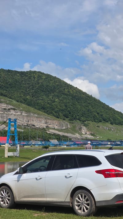 A white station wagon is parked on grassy terrain next to a body of water. In the background, a blue pedestrian bridge crosses over a river, with a lush green mountain behind it. A person wearing a light-colored cap and shirt stands on the bridge, facing away from the camera. The sky is blue with scattered white clouds, and a dark bird is visible in flight against the sky. The scene is located in the village of Tash-Asty, Russia.