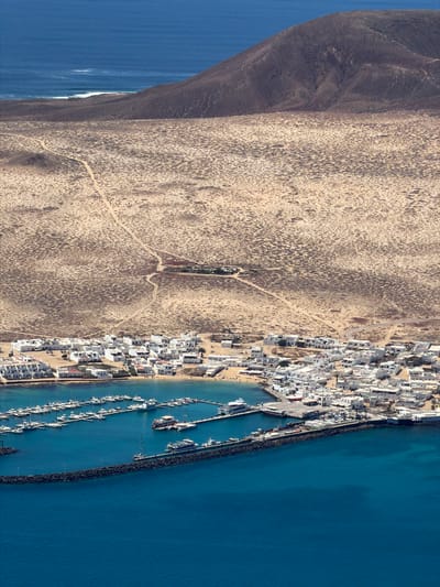 This aerial image presents a striking contrast between an arid, desert-like landscape and a bustling coastal town situated next to a vibrant blue sea. The view is oriented vertically, with the landmass on the left and the ocean on the right.

On the far left, a rugged, dark-hued mountainous area slopes down to meet a vast expanse of light brown, arid terrain, characterized by sparse vegetation or rocky textures and crisscrossed by several dirt tracks. This barren land forms a stark boundary with the urban development.

To the right of the arid landscape lies a densely built town, composed predominantly of white buildings with flat roofs, indicative of Mediterranean or Canary Island architecture. The town is concentrated along the coastline, featuring a well-developed harbor or marina. Numerous boats, ranging from small vessels to larger yachts, are visible docked at multiple piers within the sheltered waters of the harbor. A sandy beach is also visible adjacent to the town's buildings, with the clear, turquoise water lapping at its edge. A substantial breakwater, appearing as a dark, rocky barrier, extends from the coast, protecting the marina from the open sea.

Beyond the breakwater, the deep blue waters of the ocean stretch towards the right edge of the frame. Some gentle white waves are visible on the far left edge of the sea, near the more rugged coastline. The overall scene suggests a sunny day with clear weather. No individual people or visible text can be discerned due to the high aerial perspective. The image captures a dynamic coastal settlement in Haría, Spain, where human development integrates with the natural, contrasting elements of a dry landscape and the open sea.