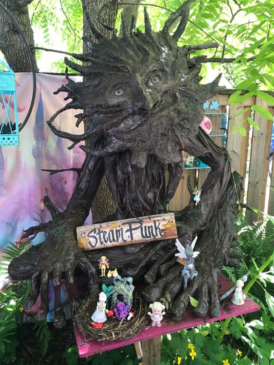 The image depicts a large, dark, sculpted tree-like figure with a face and outstretched arms, positioned outdoors under dappled sunlight filtering through green leaves. A wooden sign with "Steam Punk" written in white lettering and adorned with small yellow stars is attached to the figure's arm. Various small fairy figurines are placed around the tree sculpture. To the left of the tree figure, a turquoise decorative birdcage is visible, with a pink and purple patterned backdrop behind it. In the foreground, a nest-like structure holds several more small figurines, including dolls dressed in fairy-like attire and miniature mushrooms. Green ferns and small yellow wildflowers grow around the base of the sculpture. The background shows a wooden fence and more greenery. This scene appears to be a whimsical display at a market or event, possibly located in the city of Severn, Canada, as suggested by the context.