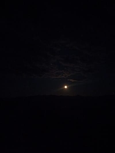 This is a vertical, dimly lit photograph taken at night in Kamyshenka, Russia. The primary subject is a bright full moon illuminating the sky, which is partially obscured by soft, scattered clouds. The moon is positioned slightly to the left of the center of the frame. Below the moon and clouds, a dark, silhouetted landscape of rolling hills or mountains stretches across the bottom third of the image. The overall scene is dark and tranquil, suggesting a calm night. The time of day is clearly nighttime, indicated by the presence of the moon and the darkness. There are no visible people, objects, or text in the photograph. The location is identified as Kamyshenka, Russia, adding a specific geographical context to the nocturnal landscape.
