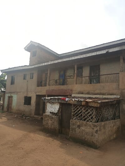 The image shows a two-story building in a residential area of Obromudu, Nigeria. The building is made of concrete blocks and has a corrugated metal roof. The building is unfinished, and several of the windows are missing glass. The building has a second floor balcony with iron railings. The building has a door on the bottom floor that appears to be unfinished, with a door in the center made of wood and a window on each side, each with a metal frame. The building is surrounded by dirt and there is a small outbuilding in front of the main building. The outbuilding is also made of concrete blocks and has a corrugated metal roof. There are also several clothes hanging on the roof of the outbuilding. The image is taken at ground level and is focused on the building and the immediate surroundings. There are no people visible in the image. The location, based on the coordinates provided, is a residential area with a dirt road and a few trees.