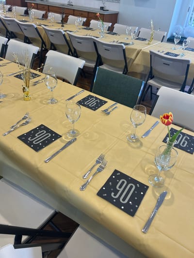 The image depicts a dining table set up for a 90th birthday celebration. The location is difficult to determine from the image; there is no clear indication this is Kelowna, Canada. A long table is covered with a light gold tablecloth. Each place setting includes a wine glass, a knife, two forks, and a black square card with "90" printed in silver. Some place settings also have a small vase with a single tulip. Other centerpieces include small gold-colored decorations. White folding chairs are arranged around the table. The background shows part of a room with white walls and some cabinetry. The lighting is bright.