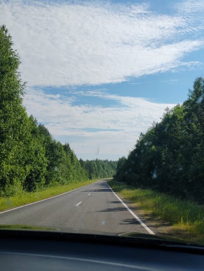 The image shows a view from a vehicle driving on a paved road through a forested area. The road has white lane markings. On both sides of the road are dense green trees. The sky above is blue with white, wispy clouds. The scene appears to be a rural or natural setting. The image is in focus and well-lit, suggesting it was taken during daylight hours. There is no indication of the specific location within Chukhlem, Russia, beyond it being a road in a wooded area.