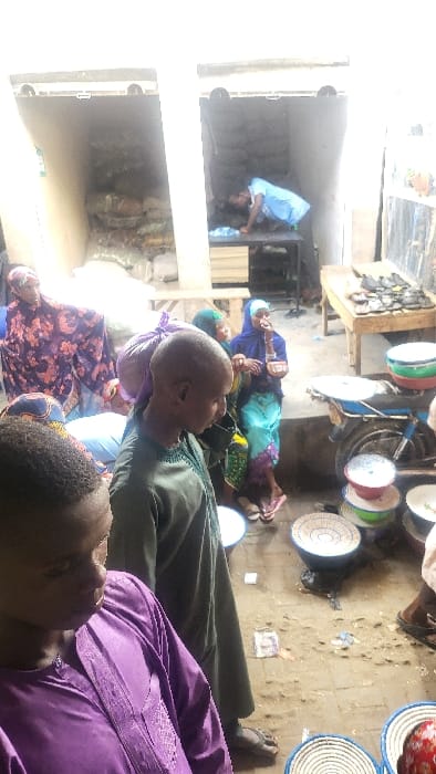 The image depicts a commercial thoroughfare or market area in Fagge D1, Nigeria. The ground surface is uneven, appearing as packed dirt or a worn paved area, with scattered debris. The scene includes approximately seven individuals and various merchandise.
In the left foreground, a young male, largely visible from the chest up, wears a purple long-sleeved garment with embroidered detail, looking down. Behind him, a male with a bald head or very short hair, wearing a green long-sleeved garment and sandals, has a purple bundle on his left shoulder.
To the immediate left mid-ground, a female in a dark purple patterned garment and head covering stands within a dim, open stall, which contains stacked bags. In the mid-ground center-right, a female wearing a light blue head covering, a patterned skirt, and flip-flops, holds a small item to her mouth as if eating. Adjacent to her is a blue motorcycle, with several white and multicolored bowls stacked on its seat and rear fender. On the ground in the foreground and mid-ground, numerous colorful, circular bowls or woven baskets are displayed.
In the background, a second dim, open stall on the right reveals a male in a light blue shirt leaning over a dark table laden with items. To the right of this stall entrance, a wooden table displays multiple dark-colored shoes or sandals. The lighting suggests an overhead light source or open-air conditions.
