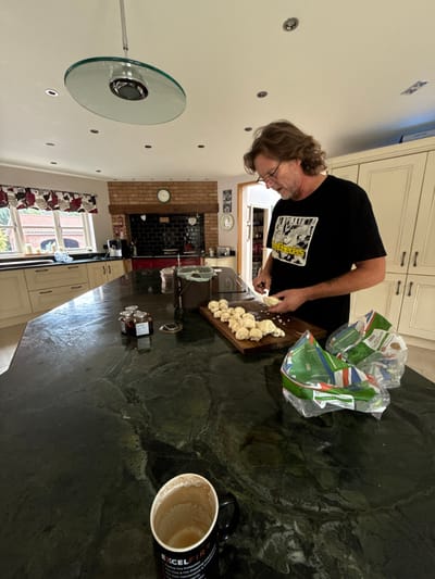 This image depicts a domestic kitchen scene in Hornsea, United Kingdom. A middle-aged male, characterized by medium-length wavy light brown hair and dark-rimmed glasses, is positioned at a large, dark green, veined granite kitchen island. He wears a black short-sleeved t-shirt featuring a white graphic, identifiable as "The Breeders" band logo. The subject is actively engaged in cutting a white vegetable floret with a small silver knife.
On the wooden cutting board directly in front of him, numerous prepared florets of what appears to be cauliflower are arranged. To the subject's right, two partially folded clear plastic bags, bearing green and white Tesco branding, rest on the countertop; one bag visibly contains additional white vegetables. In the immediate foreground, a black mug with "EXCEL FIR" text is situated on the island, containing visible residue from a beverage. Further left on the island, a cluster of small glass jars with dark lids and labels, along with a brown lidded food waste bin, are present.
The kitchen background features cream-colored base and wall cabinets with dark handles. A red range cooker is centrally located within a brick archway, set against a black tiled backsplash. A round wall clock is mounted on the brickwork above the cooker. Two large windows on the left wall, framed in white and adorned with red and white patterned curtains, offer a view of an outdoor area and a distant building. The white ceiling is fitted with multiple recessed spotlights and a prominent circular glass light fixture suspended centrally. An open doorway on the right side of the image leads to another interior space.